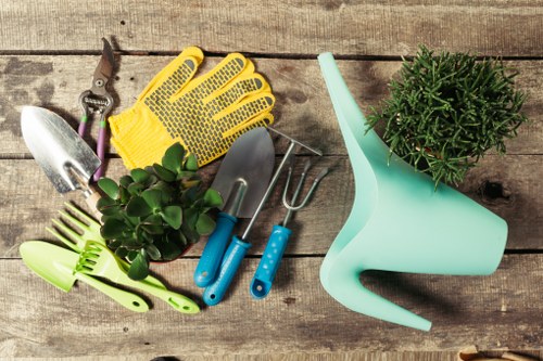 An assortment of gardening tools and supplies arranged on a weathered wooden surface, including a small pair of pruning shears, a yellow rubber glove, a trowel with a purple handle, a hand rake, a small spade with a light blue handle, a green plant sprayer, and a potted green shrub with dense foliage. The tools are clean and neatly placed, suggesting readiness for gardening tasks such as planting, pruning, or watering. The background of aged wooden planks adds a rustic outdoor feel, typical of a garden workspace at a residential property in Wandsworth. This collection of gardening essentials reflects professional garden maintenance services provided by Landscapers Wandsworth, who specialize in enhancing outdoor spaces with a focus on horticultural care and landscaping design. The arrangement emphasizes a tidy, well-organized approach to garden upkeep, suitable for front or back garden projects, and supports the overall theme of gardening and outdoor landscaping in the London area. The natural tones of the wood, metal, and plant contrast with the bright colors of the tools, creating a vibrant yet practical scene for garden service promotion.