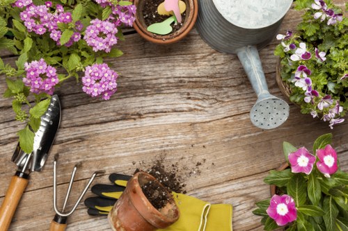 The image shows a rustic wooden garden table with various gardening tools and supplies arranged on it. There are several pots with colourful flowering plants, including pink and white petunias, positioned at different points around the table. A metal watering can with a long spout sits near the top right, while a trowel with a wooden handle is placed on the left side. Soil is spilled from a small terracotta pot, accompanied by a pair of gardening gloves in bright yellow, and a three-pronged hand fork nearby. The scene is outdoors, with natural daylight highlighting the textures of the wooden surface, soil, and vibrant plant leaves, evoking a well-maintained garden space suitable for garden renovation and outdoor landscaping services in Wandsworth.