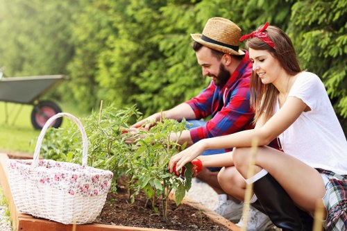 In a well-maintained backyard garden with lush green trees and shrubbery in the background, a man and a woman are actively engaged in gardening tasks. They are kneeling beside a raised flower bed or planting area, which contains young plants and soil. The man, wearing a straw hat and a red and blue plaid shirt, is using his hands to tend to the plants, while the woman, dressed in a white t-shirt and plaid skirt, also reaches into the soil, possibly planting or pruning. A white wicker basket with a floral fabric lining is placed nearby on the edge of the garden bed, containing gardening tools or supplies. The garden surface area shows a mix of soil and grass, with a tidy, organized appearance. The natural lighting suggests a sunny day, highlighting the vibrant green of the plants and the natural tones of the garden setting. The scene reflects care and engagement in outdoor gardening, aligning with landscaping and lawn care services provided by Landscapers Wandsworth, supporting a healthy, beautiful outdoor environment in the Wandsworth area.