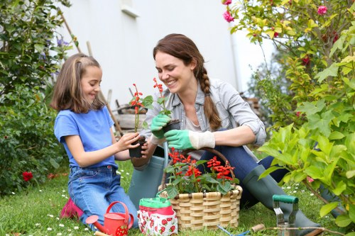 A woman and a young girl are engaging in gardening in a lush, well-maintained outdoor garden space, which features a green lawn, various flowering plants, and shrubbery. The woman, dressed in a casual grey shirt and wearing green gardening gloves, is smiling as she holds a small potted plant with red flowers, while the girl, wearing a blue t-shirt and pink shoes, is carefully examining or watering the same plant. On the ground beside them is a wicker basket filled with gardening tools and supplies, including a watering can with a floral pattern, a small red watering can, and a garden trowel. The background includes a white exterior wall, and the scene is illuminated by natural daylight, suggesting a clear weather day. The setting exemplifies a typical residential garden, highlighting outdoor gardening activities suitable for local gardening services offered in Wandsworth and the surrounding areas, with a focus on nurturing plants and maintaining outdoor greenery.