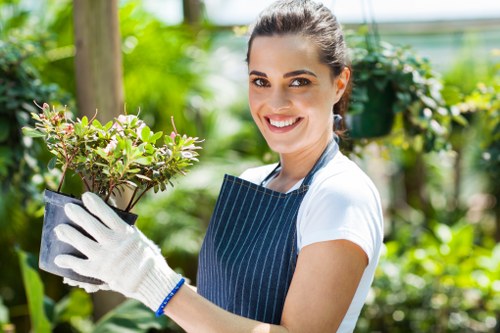 A young woman with dark hair tied back, wearing a white t-shirt, striped apron, and gardening gloves, stands in a lush garden with a variety of greenery and plants in the background. She is smiling and holding a potted shrub with small, vibrant green leaves and delicate pink flowers. The garden features neatly maintained grass, a wooden garden post, and a mix of shrubs and plants, suggesting a well-kept outdoor space typical of residential landscaping in Wandsworth. The bright, natural lighting indicates a sunny day, highlighting the healthy foliage and creating a welcoming outdoor environment. This scene reflects skilled gardening and landscaping services, emphasizing plant care, garden maintenance, and outdoor beautification, relevant to the services offered by Landscapers Wandsworth in the SW postcode area.
