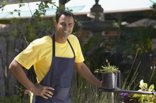 A smiling man wearing a yellow t-shirt and a dark apron is standing in a well-maintained garden, holding a black plastic pot containing a small green leafy plant. The garden features a lush lawn with dense, neatly trimmed hedges and various shrubs in the background. Behind him, there is a wooden garden fence and a decorative stone fountain or birdbath, partially obscured by foliage. The scene suggests a sunny day with natural light illuminating the outdoor space, which is typical of a residential garden in Wandsworth. This image reflects landscaping and gardening activities, highlighting the care of potted plants in an organized, leafy outdoor environment.