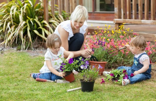 In a well-maintained front garden during daylight hours, an older woman with short, light-colored hair is seated on the grass, engaging with two young children who are planting flowers in small pots. The garden features a lush, green lawn with a variety of flower pots filled with purple, pink, and yellow blooms, indicating recent planting or gardening activity. Behind them, a vibrant flower bed brims with colourful plants, bordered by neatly trimmed shrubs and tall ornamental grasses. To the right, there is a wooden deck or patio area connected to a building with large glass doors, suggesting an indoor-outdoor living space. The weather appears bright, with natural sunlight casting soft shadows, illustrating a pleasant day suitable for outdoor gardening. The scene exemplifies a family-friendly garden setting with traditional landscaping elements common in Wandsworth, highlighting a focus on casual, accessible outdoor maintenance and local garden enhancement services by Landscapers Wandsworth.