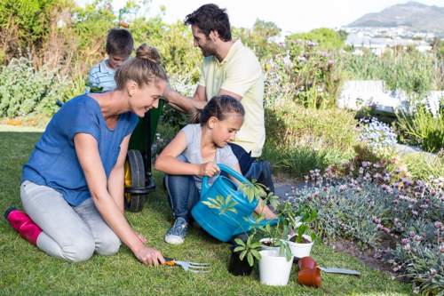 A family of four gardening in a lush backyard, with a woman planting flowers or shrubs on a well-maintained grassy lawn, while a young girl with a backpack waters or tends to plants nearby. In the background, a man and a boy are crouched down, working with garden tools amidst colorful flower beds, bushes, and hedges. The garden features a mix of lush green grass, flowering plants, and small shrubs, with a paved patio area and a scenic view of distant trees and hills under bright natural sunlight. The scene captures a moment of outdoor family activity, reflecting a well-kept, landscaped outdoor space typical of Wandsworth gardens, with a focus on planting and ground-level garden care, supported by professional gardening services like those provided by Landscapers Wandsworth.