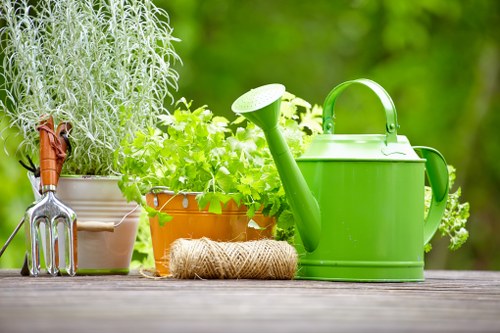 A close-up view of a garden table surface in a well-maintained outdoor space in Wandsworth, displaying a collection of gardening tools and supplies. On the left, a metal hand rake with a wooden handle and a rust-resistant fork are placed next to potted plants; the first pot contains a silver-leaved herb, possibly rosemary, and the second contains bright green, bushy foliage, likely parsley or coriander, both in terracotta and plastic pots. In front, a natural coir garden twine roll rests on the wooden table, providing a subtle hint of practical garden maintenance. To the right, a large, vibrant green plastic watering can with a curved handle and a long spout dominates the scene, indicating readiness for watering plants or lawn areas. The background features a lush garden setting with blurred green foliage and trees, consistent with a peaceful backyard or landscaped outdoor space in Wandsworth, enhancing the overall impression of professional gardening and outdoor maintenance services by Landscapers Wandsworth. The lighting suggests natural daylight, with soft sunlight illuminating the garden tools and plants, emphasizing the colour contrasts between bright greenery and neutral tones of the table surface and containers.