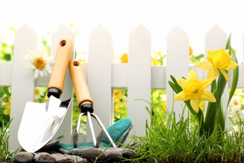 The image depicts a section of a well-maintained garden with a white picket fence in the background, situated in a residential outdoor space likely in Wandsworth. In the foreground, there is a lush, green lawn with dense grass and healthy growth, bordered by a small flower bed containing vibrant yellow flowers and white daisies. The flower bed is bordered by soil and contains a mixture of green foliage. Two gardening tools—a small hand trowel and a cultivator—are resting on the soil near the flowers, indicating recent or ongoing planting activity. The garden area is bathed in natural daylight, suggesting a bright, clear day, and the overall scene reflects a tidy, cared-for outdoor space suitable for gardening and landscaping services provided by Landscapers Wandsworth. The image emphasizes a clean, orderly garden environment with essential planting features, suitable for landscape improvement or maintenance in the local area.