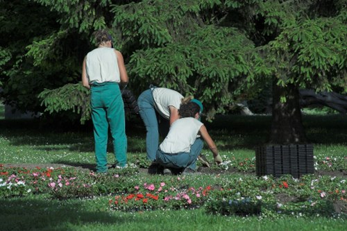 The image depicts three individuals working together in a well-maintained garden, likely in Wandsworth, with lush green trees providing shade in the background. One person is standing, wearing a sleeveless top and teal trousers, while the other two are crouched close to the ground, tending to flower beds that are edged with small, colorful blooms and dark mulch. The garden features a neatly trimmed lawn area with a variety of plants and a paved pathway or patio surface partially visible. A black plastic garden storage box is positioned on the right side of the scene. The overall setting suggests outdoor gardening activity on a clear day with natural light illuminating the vibrant greenery and floral displays, reflecting professional landscaping and lawn care practices relevant to local outdoor spaces managed by companies such as Landscapers Wandsworth.