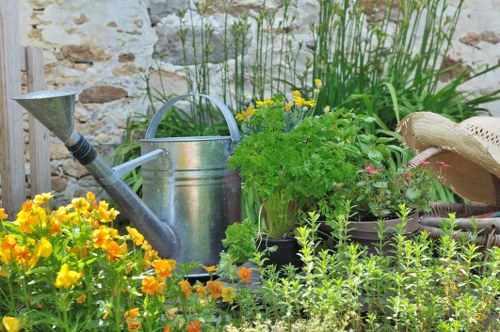 A vibrant garden scene featuring a variety of plants and gardening tools set against a rustic stone wall. In the foreground, there are bright yellow and orange flowering plants with lush green foliage, providing a splash of colour. Behind these, taller green plants with narrow leaves grow towards the sky, creating vertical interest. To the right, a metal watering can with a curved handle and spout is placed among the plants, indicating outdoor gardening activity. Nearby, a straw sun hat rests on a surface, suggesting the presence of a gardener. The garden layout includes a mix of flower beds and dense greenery, with a compost or soil bed visible in the background. The lighting appears natural, suggesting a clear or partly cloudy day. This outdoor space offers a colourful and functional garden area typical of Wandsworth, maintained by professional local gardeners. The scene exemplifies a well-tended garden with a combination of flowering plants and green foliage, suitable for outdoor gardening services to enhance or maintain.
