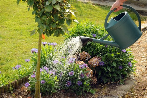A person watering a flowerbed in a well-maintained garden using a dark green watering can. The garden features a variety of plants, including a young tree with a wooden stake for support, purple and white flowering plants, and lush green shrubs. The flowerbed is edged with small wooden fencing, and the surrounding area includes a neatly cut grassy lawn. In the background, there are additional plants and a paved pathway, indicating a landscaped outdoor space typical of residential gardens in Wandsworth. The scene is illuminated by natural daylight, suggesting a clear, sunny day, and reflects professional gardening practices in a landscaped garden setting. This setting demonstrates outdoor maintenance work done by landscapers Wandsworth, focusing on plant care and garden irrigation.