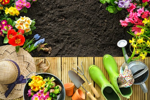 A rectangular outdoor garden space with a dark, freshly turned soil area at the top, bordered by a wooden decking section at the bottom. The garden features colorful flowering plants along the top and left edges, including pink, yellow, red, and purple blossoms, adding vibrancy to the scene. On the decking, there are various gardening tools and equipment, such as a small trowel, a hand rake, and orange plant pots, alongside bright green rubber gardening boots and a watering can. A wide-brimmed straw sun hat with a purple ribbon sits on the left side of the decking, near a potted plant with yellow and pink flowers, suggesting recent gardening activity. The environment appears well-maintained, suitable for engaging in gardening and outdoor landscaping, with natural light indicating a clear weather day, possibly in Wandsworth or nearby areas in London. The scene captures an organized outdoor gardening space, ready for planting or ongoing maintenance, reflecting professional landscaping and garden care services.