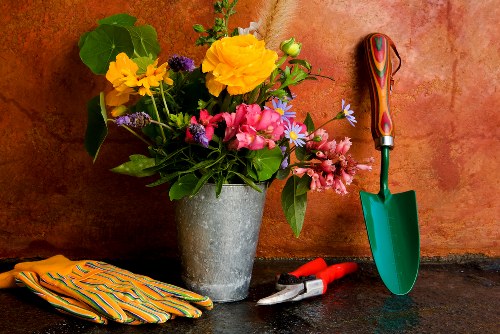 A close-up view of a gardening workspace featuring a large grey flowerpot filled with a vibrant assortment of flowers, including yellow, pink, purple, and blue blooms, arranged against a warm, textured terracotta wall. To the right of the flowerpot, there is a small green-handled trowel with a red and orange striped handle, standing upright on a dark, weathered surface. Next to the trowel, a pair of red pruning shears rest on the work surface, and to the left, a pair of yellow and orange striped gardening gloves is neatly placed. The scene captures the tools and floral elements typically associated with outdoor gardening activities in a residential garden setting, emphasizing natural tones and practical equipment used in maintaining flower beds and plant arrangements, consistent with landscaping and gardening services in the Wandsworth area.