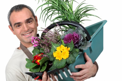A professional gardener in Wandsworth holding a blue plastic garden tool filled with a variety of vibrant flowering plants, including yellow, purple, and red blossoms, with fresh green foliage. The background shows a well-maintained outdoor space with a lush lawn, bordered by a hedge or shrubbery, indicating an organized garden layout. The area appears to be outdoors on a clear day, with natural lighting enhancing the natural tones of the soil, plants, and grass, suggesting a recent or ongoing flower bed creation or planting service. This scene reflects typical landscaping and garden maintenance activities offered by companies like Landscapers Wandsworth, focusing on creating attractive flower beds in residential gardens within the Wandsworth area.