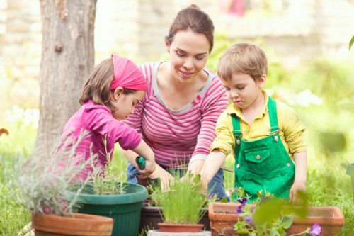 A woman with dark hair tied back, wearing a pink and white striped long-sleeve shirt, is outdoors in a garden setting during daytime. She is engaging with two young children; one girl in a pink headband and purple top, and a boy in a yellow shirt with green overalls, both actively involved in gardening activities. They are leaning over a variety of potted plants, including small flowering plants and herbs, placed on a garden table or surface among other plant containers. The background features green foliage, a tree trunk, and blurred yellowish structures, suggesting a backyard or residential garden environment. The scene is illuminated by natural light, indicating a clear or partly cloudy day, and demonstrates a moment of outdoor gardening or planting activity, aligning with landscaping and garden maintenance services provided by Landscapers Wandsworth, focusing on creating vibrant, well-maintained outdoor spaces in Wandsworth or nearby areas like SW17.
