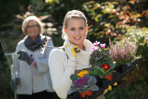 A young woman in a white sweater and yellow gardening gloves holds a black tray filled with colorful flowering plants, including red, purple, and yellow blooms, in a well-maintained garden. The garden features a lush green lawn with dense foliage and trees in the background, bathed in natural sunlight. Behind her, an older woman with glasses, wearing a white jacket and a dark scarf, is smiling and holding gardening shears, indicating that maintenance or planting work is ongoing. The scene suggests a typical outdoor gardening or landscaping activity, highlighting plant preparation and garden care in a backyard or landscaped outdoor space within Wandsworth, London, with clear seasonal daylight conditions. This setting may be associated with professional gardening services offered by Landscapers Wandsworth, demonstrating plant handling and garden beautification in a quiet residential garden environment.