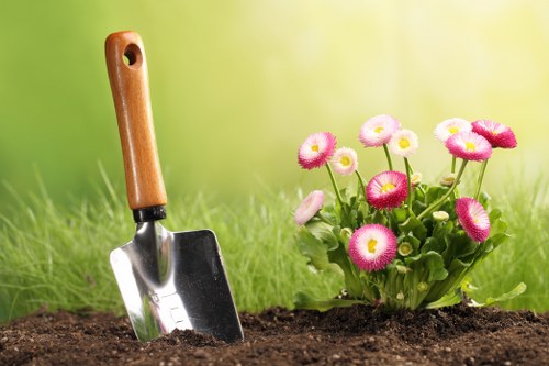 A close-up of a garden scene with freshly dug, dark brown soil in the foreground, featuring a small hand trowel with a wooden handle partially embedded into the soil. To the right, there is a vibrant cluster of pink and white flowering plants with rounded blooms and green foliage, suggesting a well-maintained flower bed. The background consists of blurred green grass and a natural outdoor setting, indicating a sunny day with bright, natural lighting. This scene exemplifies typical gardening activities that might be provided by landscapers in Wandsworth, including planting and flower bed maintenance, situated within a neat and organized outdoor space in a residential or community garden environment.