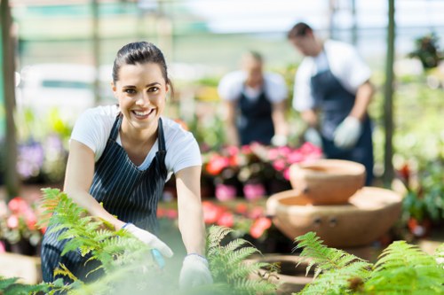 In a well-lit greenhouse environment, a woman with dark hair tied back is smiling at the camera while tending to lush green potted plants, including ferns, in a garden centre or nursery setting. She is wearing a striped apron over a white shirt and appears to be engaged in planting or watering activities. Behind her, blurred figures of other garden staff or customers are visible, also working among vibrant flowering plants and decorative water features, such as a stone fountain. The greenhouse has large glass panels that allow natural light to illuminate the space, highlighting the variety of greenery and floral displays. The scene reflects a professional gardening or landscaping service environment, with a focus on maintaining healthy plants and outdoor landscaping elements suitable for residential gardens in Wandsworth or the surrounding area. The overall atmosphere is bright, clean, and serene, emphasizing skilled plant care and outdoor horticultural work.
